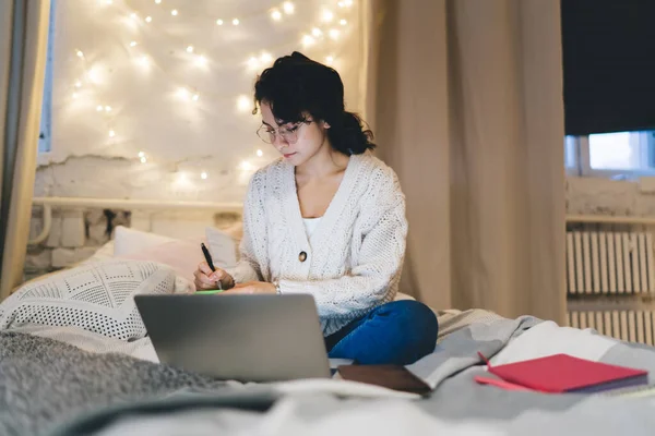 穿着毛衣和眼镜的女自由撰稿人在笔记本电脑上工作时坐在舒适的床上 并在抄本上记笔记 — 图库照片