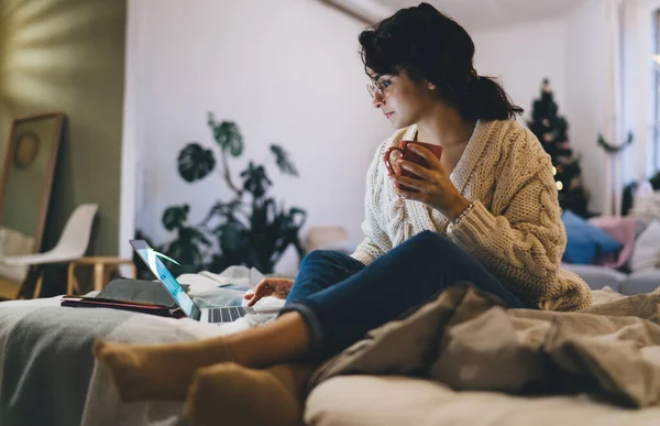 Low angle of focused woman in eyeglasses and casual clothes sitting on comfortable bed and drinking cup of coffee during work on laptop