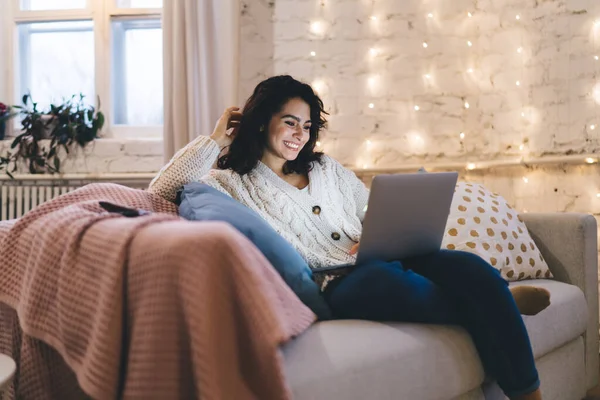 穿着保暖衣服的漂亮女性坐在客厅舒适的沙发上 一边微笑着一边浏览笔记本电脑 — 图库照片