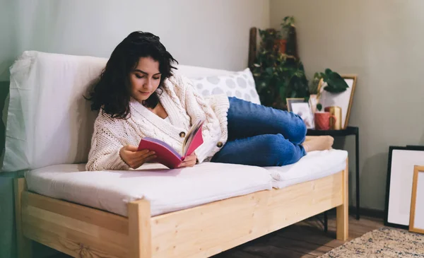 Full body concentrated female in warm clothes lying on side on comfortable couch while checking plans on day reading notebook against interior objects near gray wall