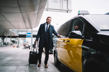 30 yaşındaki neşeli beyaz işadamının tam boy portresi havaalanından taksi transferine yakın poz veren başarılı erkek gururlu CEO valiziyle taksi ulaşımının yanındaki kameraya gülümsüyor.