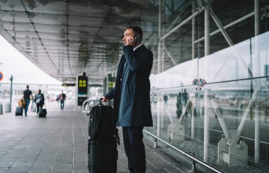 Beyaz iş adamı havaalanı terminalinin yanında bekliyor. Şirket ortağıyla uluslararası bir konuşma yapmak için cep telefonu kullanıyor. Resmi giyinmiş patron iş seyahatleri sırasında cep telefonuyla iletişim kuruyor.