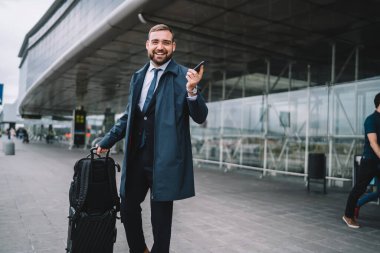 Bavul taşıyan neşeli iş adamının yarım boy portresi kameraya gülümserken taksi siparişi veren başarılı erkek girişimci hakkında akıllı telefon doğrulama bültenini bekliyor.