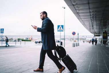 Kafkasyalı yolcu, havaalanının dış cephesinde modern cep telefonu cihazıyla elektronik bileti kontrol ediyor. Resmi olarak giyinmiş, bagaj taksisi sipariş eden bir iş adamı.