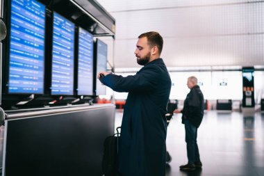 Siyah ceketli şık bir erkek, varış ve kalkış monitörünün yanında duruyor ve havaalanındaki kol saatini kontrol ediyor.
