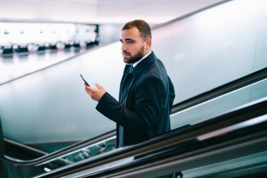 Modern mobil teknolojiye sahip düşünceli bir erkek yolcu uçuş bilgilerini düşünürken elektronik merdivenlerden havaalanı terminaline iniyor, yürüyen merdivende akıllı telefon cihazıyla tefekkür eden bir işadamı.