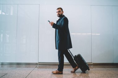Nakil servisinden e-posta bekleyen valizli ciddi iş adamının tam boy portresi. Modern cep telefonlu Kafkas işadamı havaalanında yürürken kameraya bakıyor.