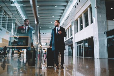 Bavul ve akıllı telefon teknolojisiyle tefekkür eden şirket yöneticisi havaalanındaki terminal kapısını ararken düşünceli, iş gezisi sırasında 30 'lu yaşlarda beyaz işveren.