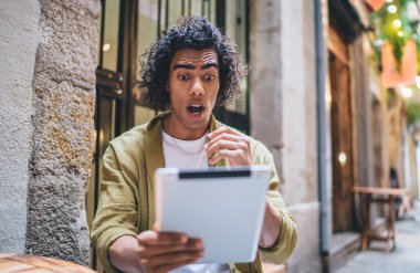 Şaşırmış kıvırcık hippi adam dijital tablet üzerinde video izliyor web sayfasında boş zaman geçiriyor, duygusal genç adam sosyal medyadan son haberleri okuyor touchpad tutuyor.