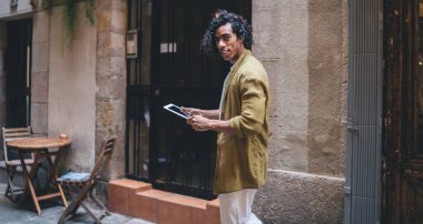 Şehir caddesindeki sokak kafesinin terasında dikilip kameraya bakarken tablet kullanan, günlük kıyafetleri içinde tatmin olmuş etnik bir erkek.