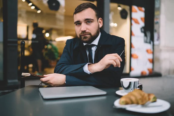 Half length portrait of formally dressed male owner sitting at cafeteria table with closed laptop device and looking at camera while waiting business partner for breakfast, Caucasian boss posing