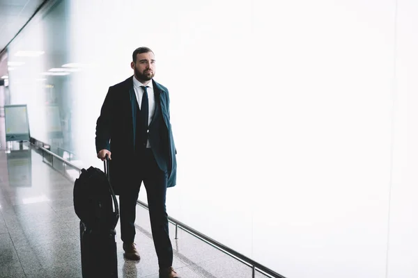 正式なスーツ姿の髭のハンサムな男は スーツケースを運び 空港ビルの白い輝く壁に対して空港廊下を歩いています — ストック写真