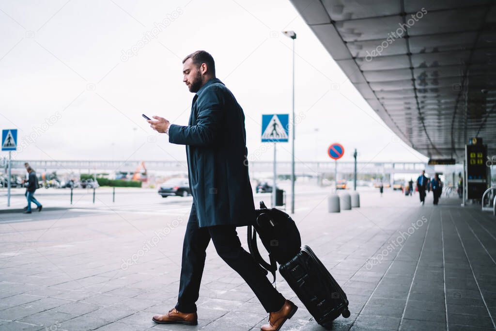 Caucasian passenger checking electronic ticket with info about terminal departure using modern cellphone gadget at airport exterior, formally dressed businessman with baggage ordering taxi cab