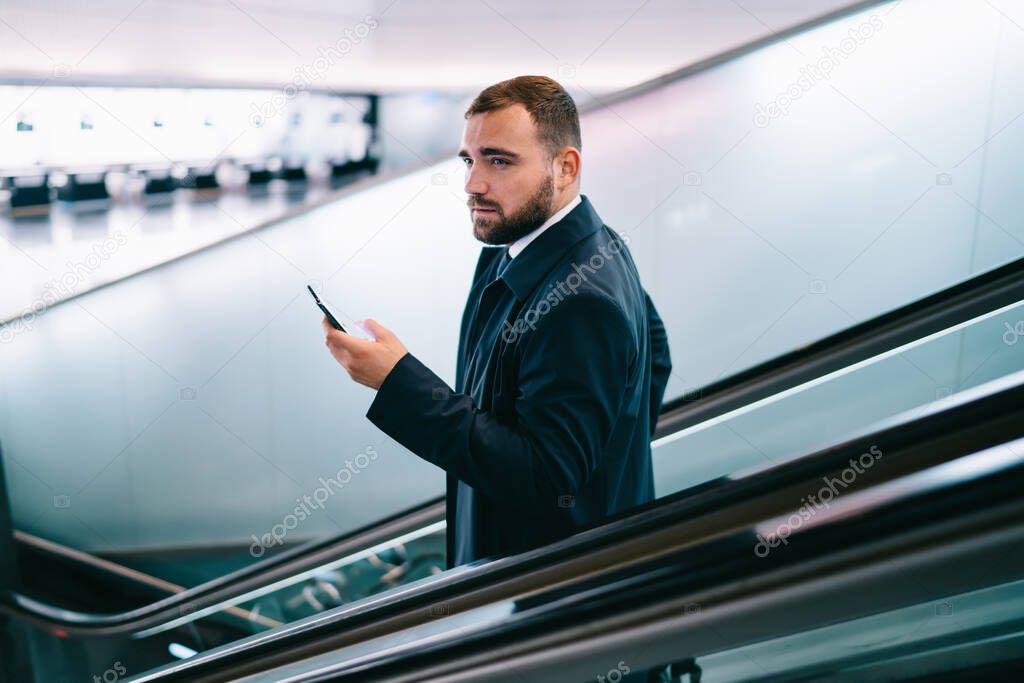 Pensive male passenger with modern mobile technology in hand pondering on flight info go down on electronic stairs to airport terminal, contemplative businessman with smartphone gadget at escalator