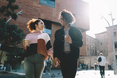 Modern binalara karşı meydanda yürürken el ele tutuşup kahve içen ve birbirlerine bakan romantik çiftlerin alçak açısı.