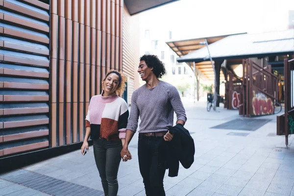 Modern binanın yakınında yürürken gülen ve konuşan sıradan giyinen mutlu siyah çift.