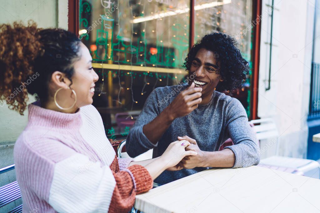Happy young multiracial couple in casual clothes laughing holding hands and looking to each other while chilling in outdoor cafe in daytime
