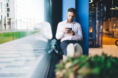Beyaz erkek blogcu pencere kenarında oturuyor ve modern hücresel teknolojide 4G kablosuz bağlantı kullanıyor, SMS mesajı göndermek ve yayınlamak için yakışıklı milenyum içerik metni