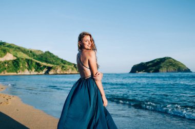 Uzun mavi elbiseli, neşeli sarışın bir kadının, parlak mavi deniz kıyısında ve yeşil adalarda huzurlu kumsalda gezinirken etrafını dönüşünün yan görüntüsü.