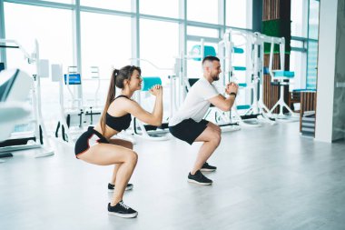 Aktif giyimli neşeli genç sporcuların ferah spor salonunda çömelme egzersizi yapıp gözlerini çevirmelerinin yan görüntüsü.