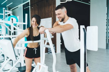 Genç ve neşeli bayan spor giyim eğitmeni spor salonunda genç ve formda bir adamın antrenman yaparken modern spor simülatörünün ekranına tıklıyor.