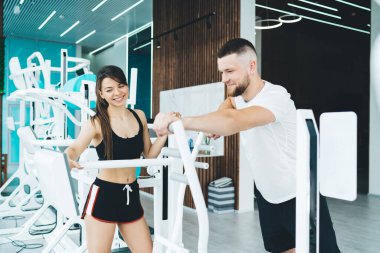 Genç gülen antrenör spor salonunda modern simülatör kullanıyor ve adam spor salonunda LED ışıklarla egzersiz yaparken ekranda tıklıyor.