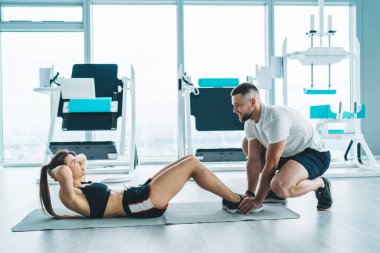 Aktif giyimli, uzun boylu, kararlı bir sporcunun spor salonunda antrenörle birlikte spor minderi üzerinde oturması.