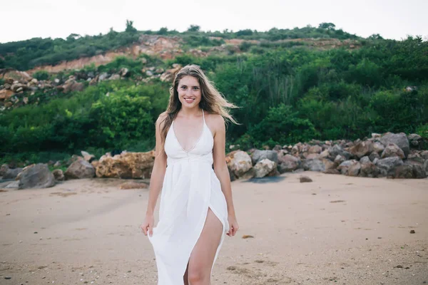 Charming delighted woman in long white sundress smiling and looking at camera while strolling along idyllic sandy shore at sunny day