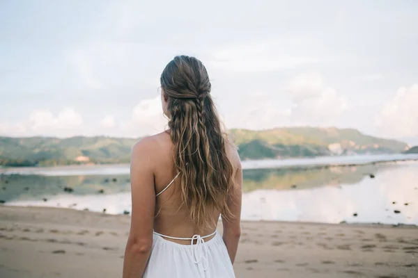 From behind blonde woman with long hair in white sundress contemplating and strolling along idyllic sandy shore at sunny day