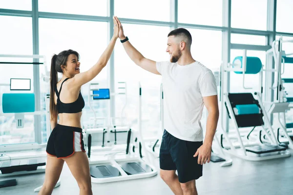 Cheerful fit sportspeople in activewear standing in modern gym and giving high five while smiling and looking at each other