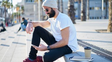 Gündüz gözlüklü, şapkalı, güneş gözlüklü, merdivenlerde oturan, cep telefonunda mesaj yazan ve güneşli bir günde boş zamanın tadını çıkaran genç hippi erkek.