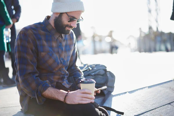 Crop smiling bearded male in casual wear hat and sunglasses typing text message on mobile phone and enjoying takeaway coffee while sitting on step in city street on sunny day
