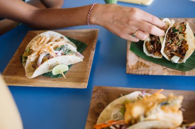 İnce bileziği ve yüzüğü olan, zarif ve zarif bir bayan mavi masadaki dikdörtgen tabaklarda servis edilen farklı takoları yiyor.