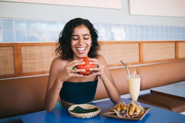 Genç iyimser etnik bayan, biftek köfteli iştah açıcı burger yerken bir tabak taco ve taze meyve püresiyle masada otururken eğleniyor.