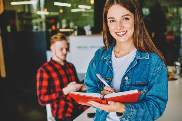 Половинный портрет жизнерадостного кавказского студента, делающего журналистский проект с использованием ручки и учебника, успешной женщины с учебником знаний в руках, улыбающейся в камеру