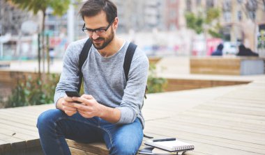 Pozitif erkek girişimci cep telefonu kullanıyor ve defter yakınındaki ahşap bankta otururken dışarıda yeni bir başlangıç konsepti üzerinde çalışıyor.
