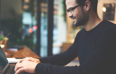 Tahta masada otururken günlük giysiler ve gözlüklerle çalışan pozitif sakallı erkek girişimcinin mahsül manzarası.