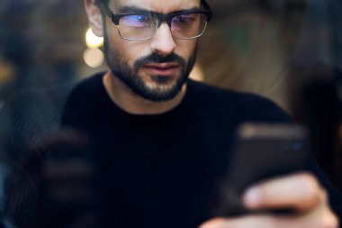 Kafede oturan ve cep telefonlarını karıştıran sıradan giysili ve gözlüklü ciddi sakallı bir adamın penceresinden.