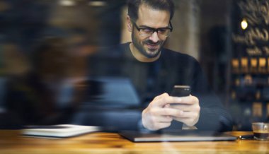 Sıradan giyinmiş, gözlüklü, kafeteryada oturan ve akıllı telefondan mesajlaşan pozitif sakallı bir erkeğin penceresinden.