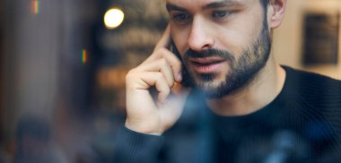 Pencere düşük açılı, sakallı, ciddi, sıradan kıyafetler giyen, kafede otururken cep telefonuyla konuşan...