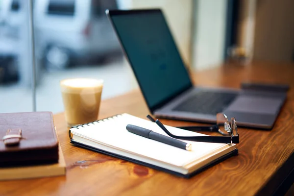 Workplace with notepad eyeglasses and pen placed on wooden table with opened laptop and glass of latte against blurred city street