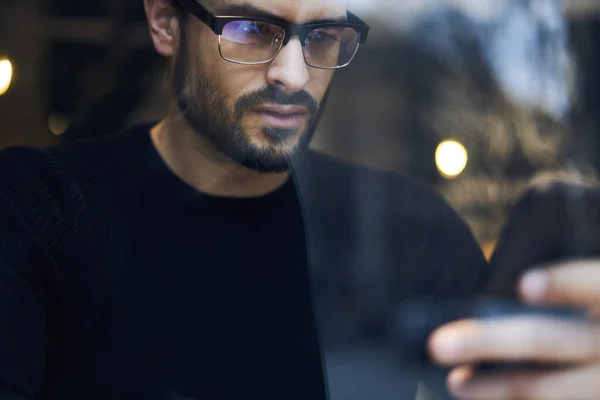 Through window of crop ethnic focused bearded male in glasses and casual outfit using mobile phone while resting in cafe