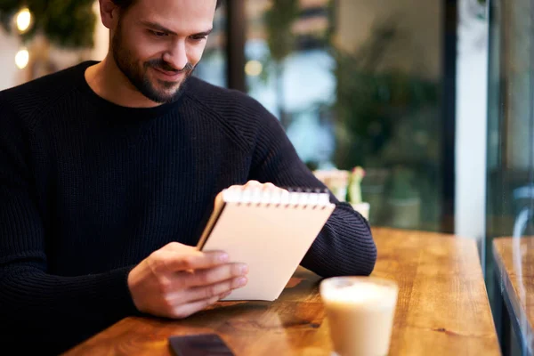 穿着休闲装的满脸胡须的男性坐在现代浅色咖啡馆里 面带微笑 并在笔记本上写着便条 — 图库照片