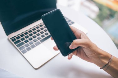 Yukarıdan gelen isimsiz erkek girişimci, proje sırasında ahşap masada dizüstü bilgisayarla oturuyor ve cep telefonuna bakıyor.