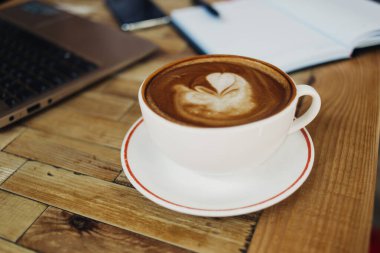 Modern, rahat bir kafede dizüstü bilgisayarın ve defterin yanındaki ahşap masaya yerleştirilmiş beyaz kapuçino bardağının üstünden.