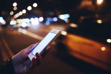 Siyah ceketli, elinde dikey pozisyonda cep telefonu ve mesajlaşan, akşam vakti parlayan Bokeh ışıklarının yanında duran kimliği belirsiz bir kadının yan görüntüsü.