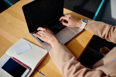 Yukardan tanınmayan, akıllı saati olan, modern dizüstü bilgisayarını karıştıran, tahta masada elinde kalem ve akıllı telefonuyla oturan tanınmamış bir kadından.