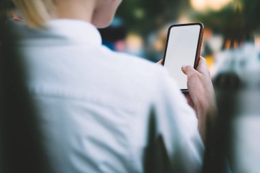 Arka planda, kısa sarı saçlı, beyaz gömlekli, güneşli bir günde sokakta duran, sörf yapan bir kadın var.