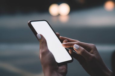 Manikürlü elleri olan tanınmayan bir kadın akşam vakti bulanık arka planda sokakta dikilirken boş ekranla modern akıllı telefona göz atıyor.