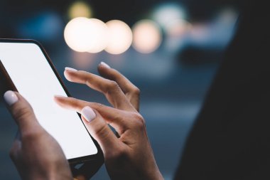 Siyah kıyafetli, karanlıkta aydınlık sokakta dururken cep telefonuna dokunan kimliği belirsiz bir kadının yan görüntüsü.